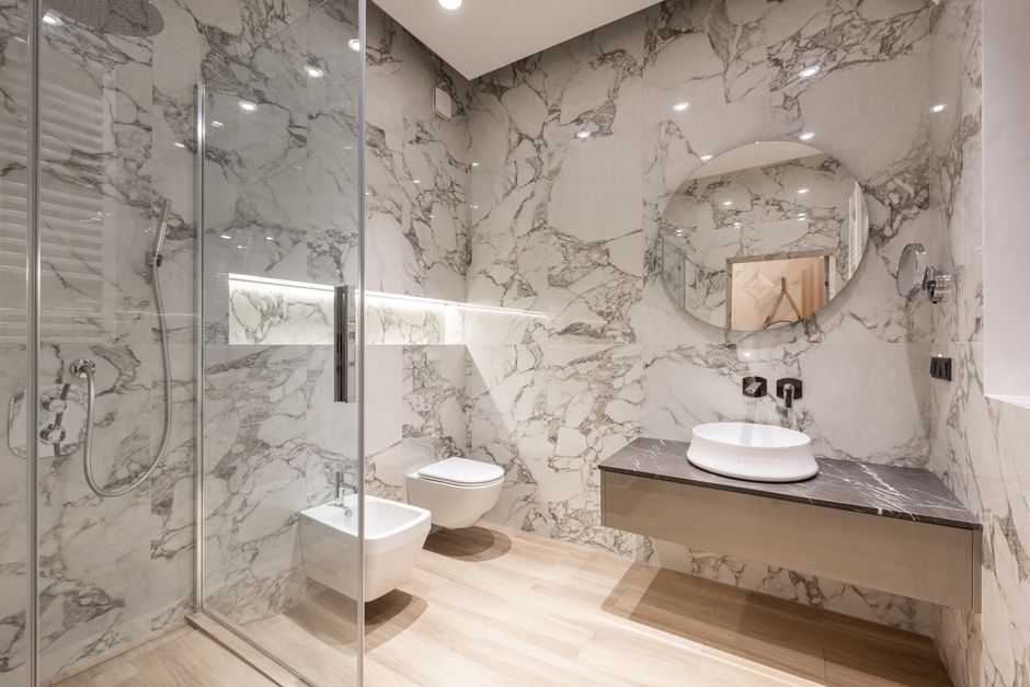 Interior of modern bright bathroom with bidet and toilet near shower and sink on counter under round mirror on tile